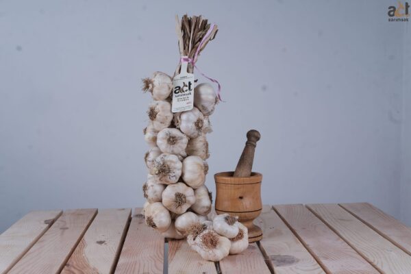 Bir demet Hediyelik Örgü Taşköprü Sarımsağı (3KG) ahşap bir masa üzerinde havan ve havaneli yanında etiketiyle birlikte dik durmakta, önünde birkaç gevşek sarımsak soğanı bulunmaktadır. Arka plan sade ve açık renklidir.
