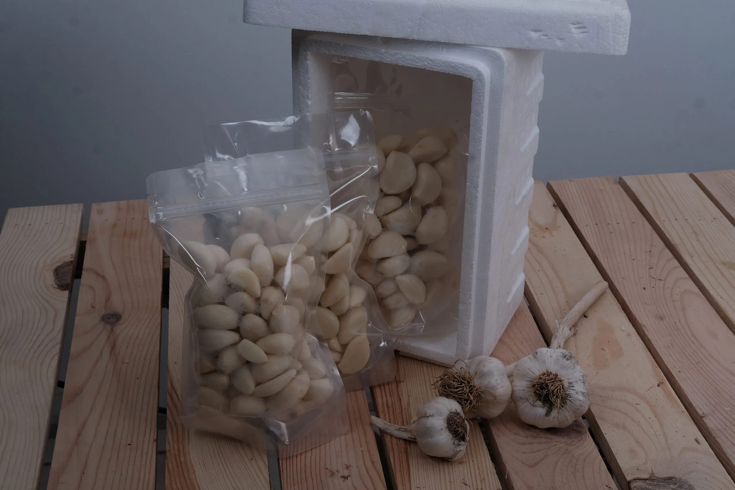 İki torba Soyulmuş Sarımsak 1000 gr soyulmuş sarımsak beyaz bir strafor kabın yanında duruyor, üç bütün sarımsak soğanı da yakınlardaki ahşap bir yüzeye yerleştirilmiş.