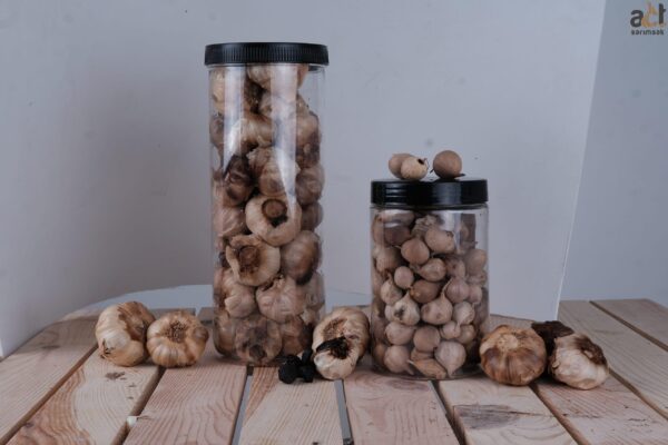 Siyah kapaklı iki şeffaf kavanoz Siyah Sarımsak (200GR) ahşap bir yüzeye yerleştirilmiş, etrafı gevşek sarımsak soğanları ve dişleriyle çevrelenmiştir. Arka plan düz beyazdır.
