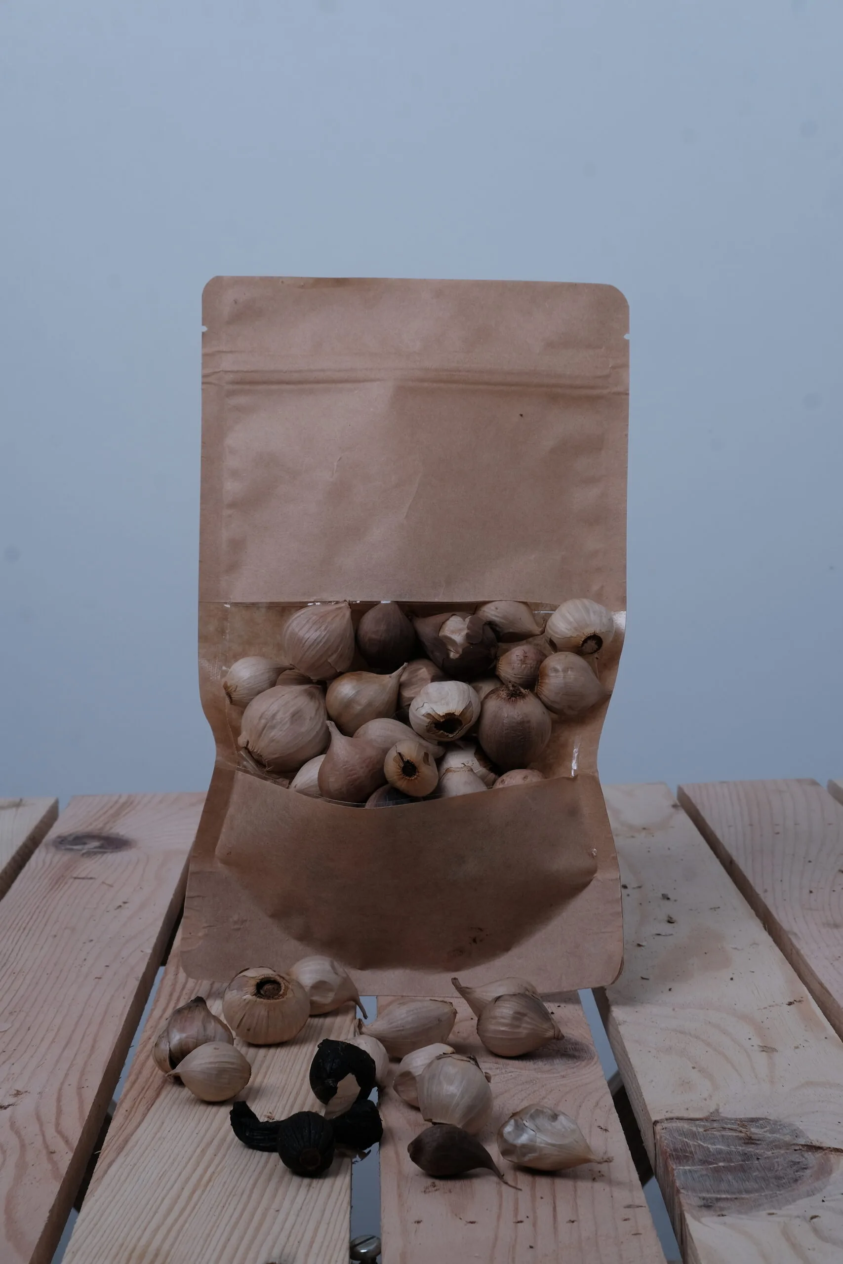 Açılıp kapanabilir kahverengi bir Siyah Sarımsak (200GR) torbası, önünde birkaç kastamonu sarımsağı soğanı ve soyulmuş bir diş siyah sarımsakla birlikte ahşap üzerinde dik durmaktadır.