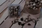 İçinde bütün siyah sarımsak soğanları bulunan 200 gramlık Siyah Sarımsak kabı ahşap bir zeminde duruyor, yanında birkaç soyulmuş diş ve gevşek soğanlar var.